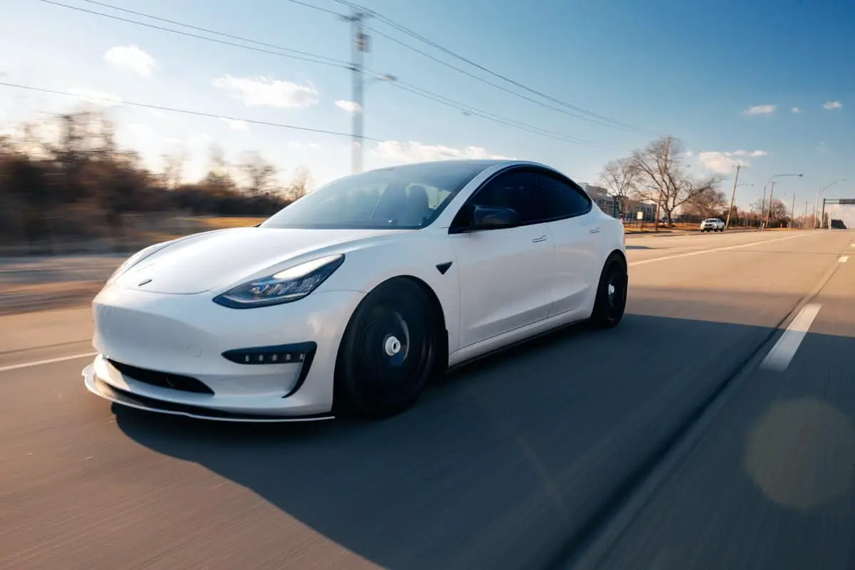 photo of a white tesla electric vehicle car driving along a street with background motion blur and blue sky