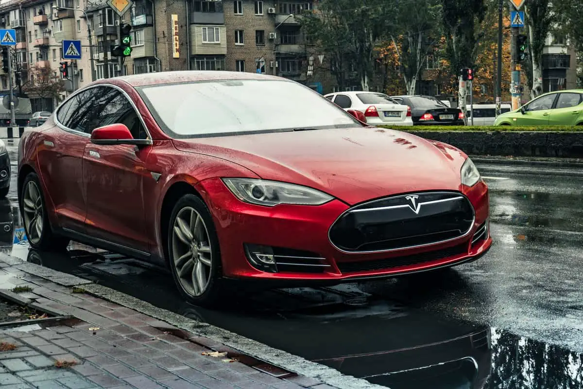 a photo of a metallic red tesla electric vehicle car parked on a street in rainy weather