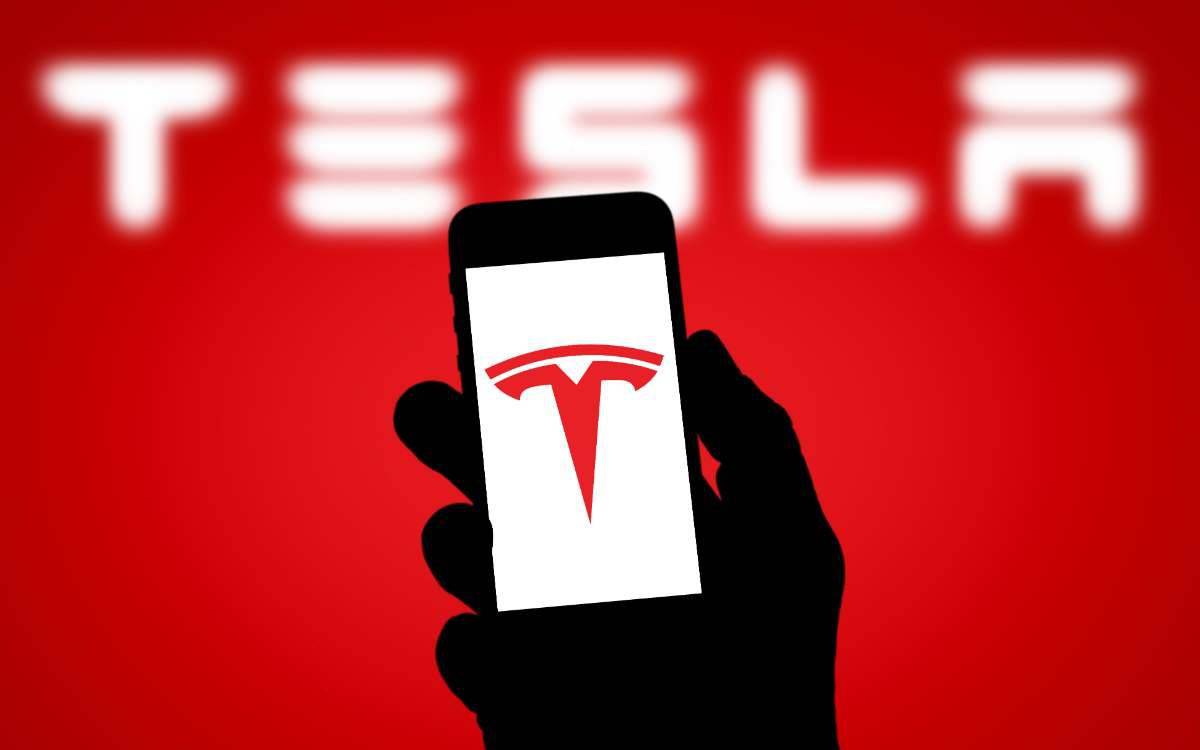 cell phone held in hand silhouetted showing tesla logo on cell phone screen with tesla logo in background
