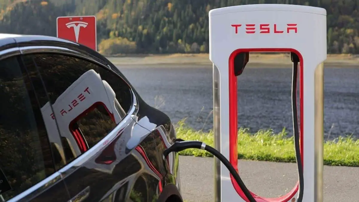 photo of a tesla fully electric vehicle being charged by a tesla supercharger on a road beside a lake in a mountainous region with forest